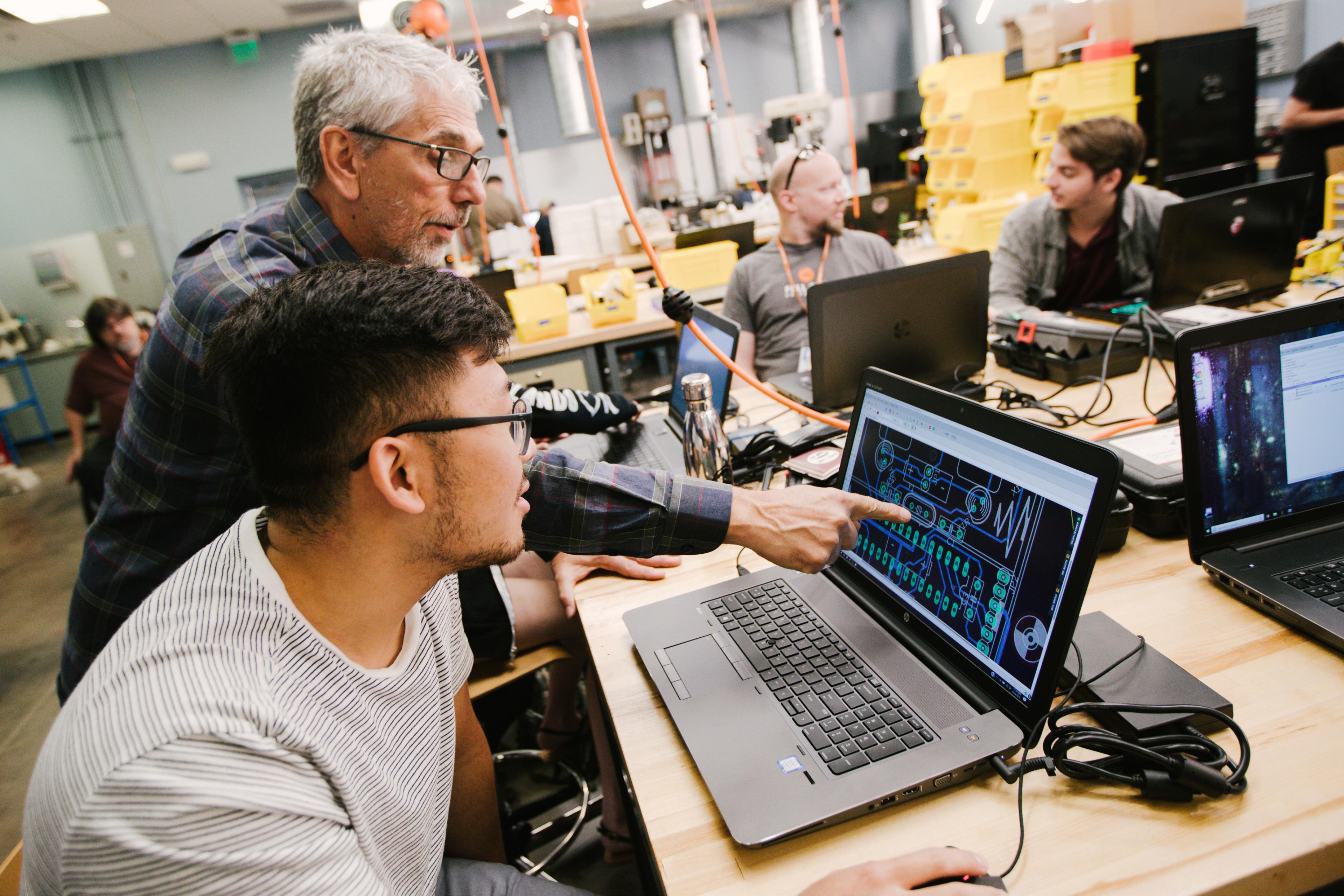 Inside the Fab Lab, an instructor points to a schematic on a screen in front of a student while other students chat in the background.
